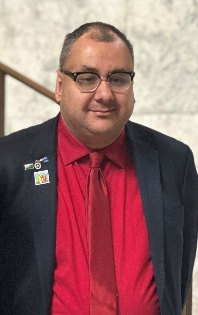 An image of Advocacy Manager John Lemus. John is a self-advocate. He is wearing a red shirt with a red tie, and a black blazer. He has the Best Buddies pin on his lapel. He is also wearing glasses.