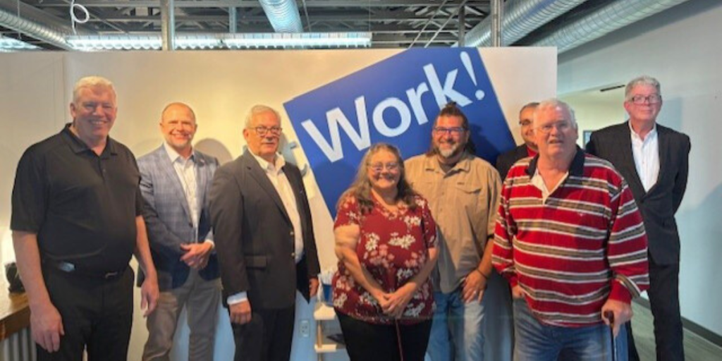 A group of people from AtWork!, Source America, and AbilityOne standing in the front lobby of AtWork!, with the AtWork! logo in the background.