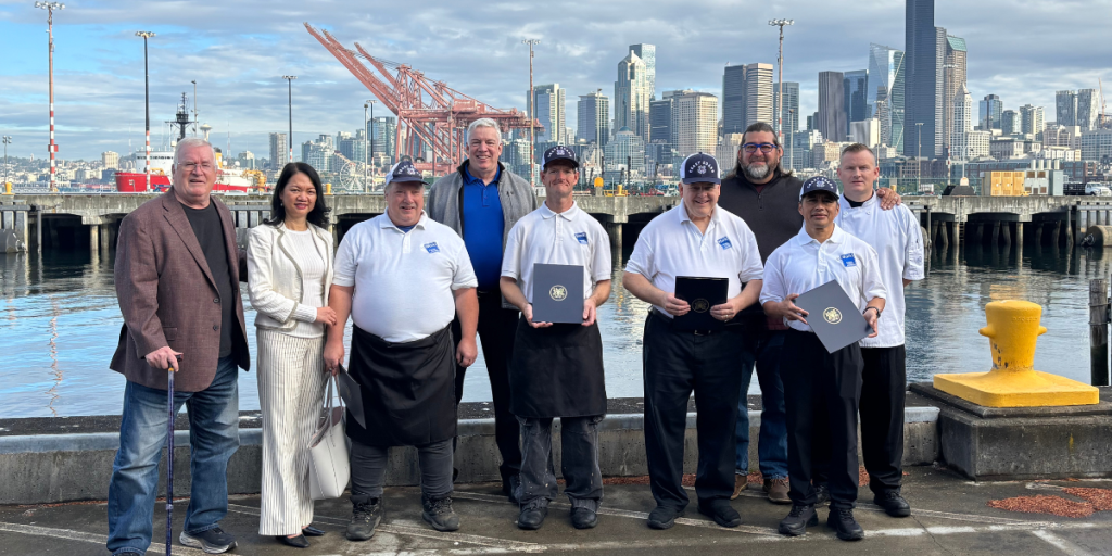 The BPAW Coast Guard Food Services team and AtWork! leadership stand in front of the Seattle Skyline with their recognitions.