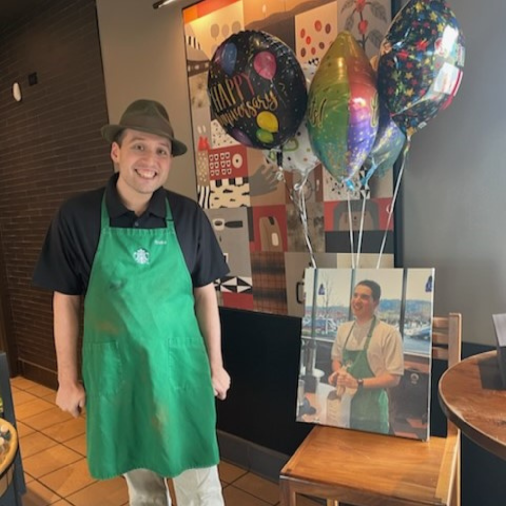 Blake celebrates 20 years with Starbucks. He is wearing a green apron and a gray fedora. He is standing next to a photo and balloon display.