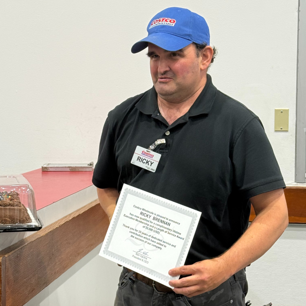 Ricky, wearing a blue Costco Hat, a black polo, and his new Silver Badge. He is holding his certificate from Costco, celebrating his 25 years with the company.