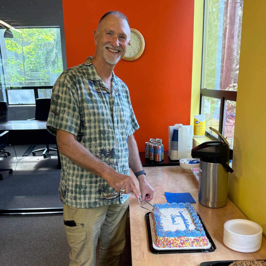 Steve Repanich cuts his retirement cake. He is wearing a green checkered shirt and khaki pants.