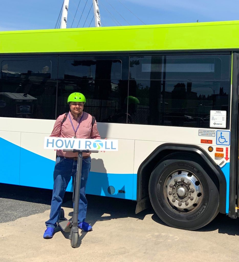 Advocacy Manager John Lemus poses next to a city bus with his scooter. He is wearing a green helmet, pink shirt, and blue jeans.